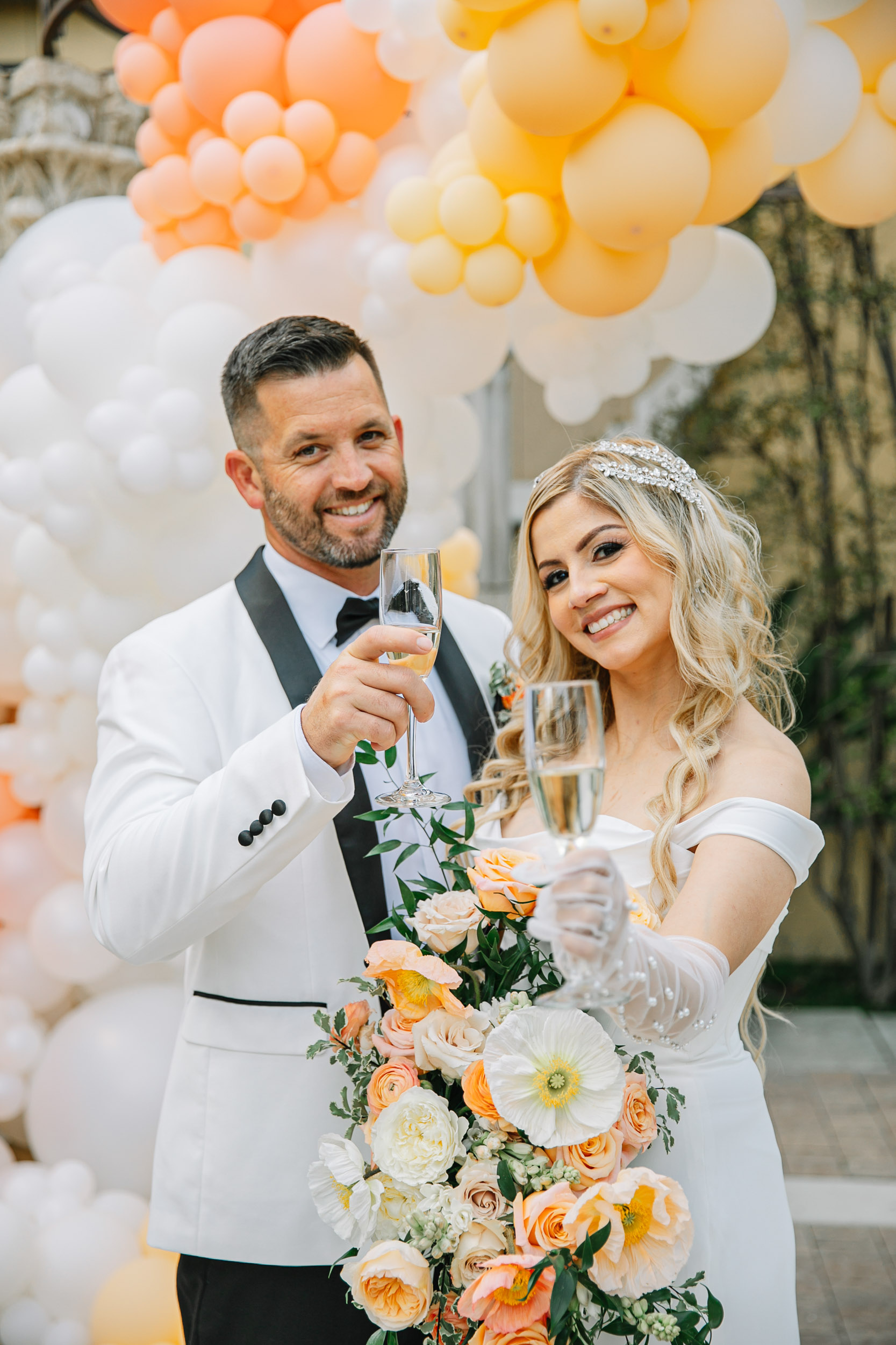 Bakersfield wedding couple with champagne glasses and a bridal bouquet of soft yellows and orange poppy flowers and garden roses pose standing in front of a balloon garland.