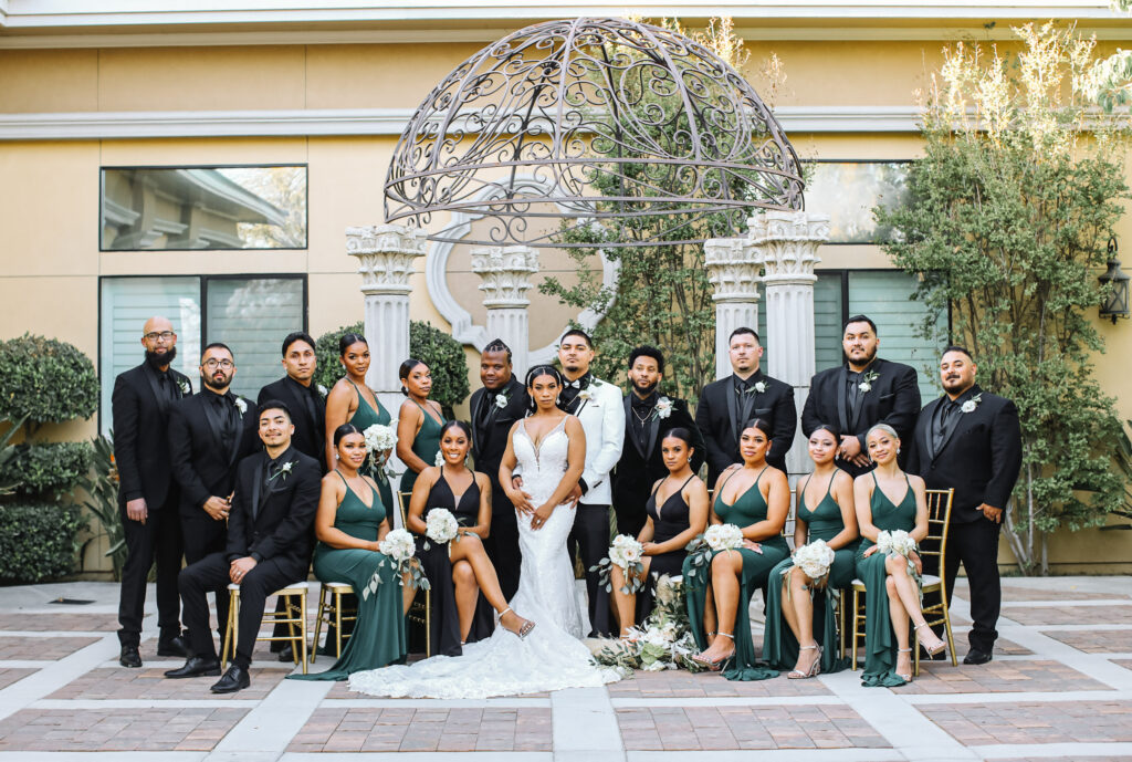 Wedding party in black and emerald green posing with the couple in the garden courtyard of this tuscan inspired Bakersfield venue.