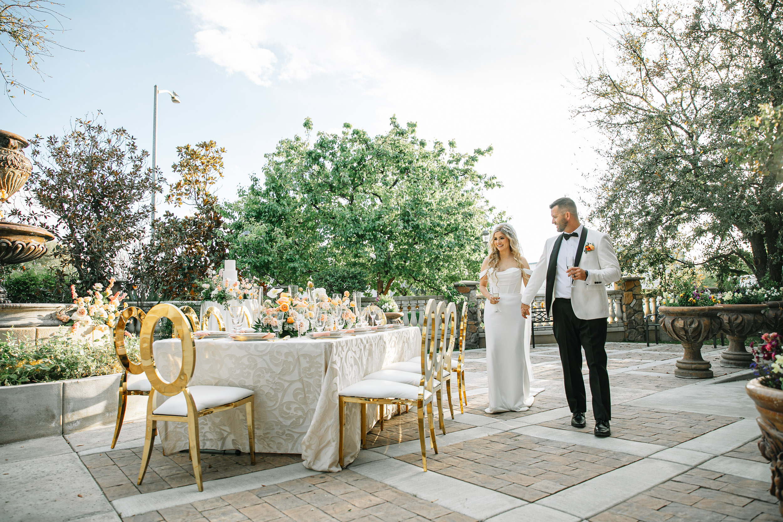Bride in a modern wedding dress and groom in and black and white tuxedo stand in the garden courtyard of a Bakersfield wedding venue admiring a beautiful spring tablescape that includes ivory, peach and pink flowers, candles, and gold chairs.
