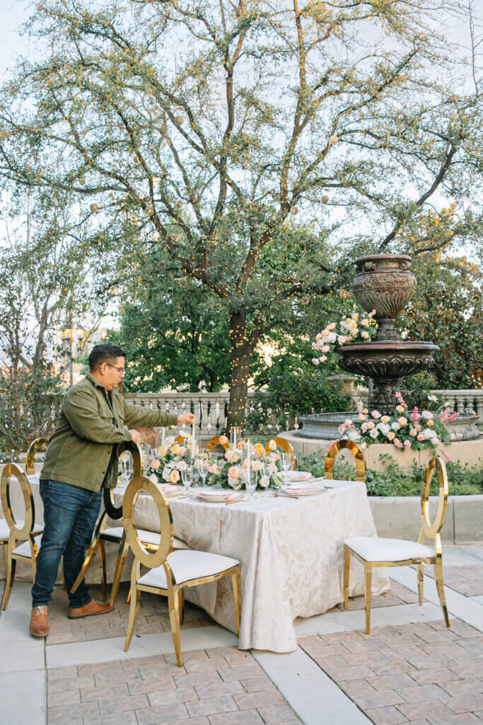 Carlos of Make It Happen Events sets up a wedding table for a styled shoot taking place in the garden courtyard of an elegant indoor wedding venue.