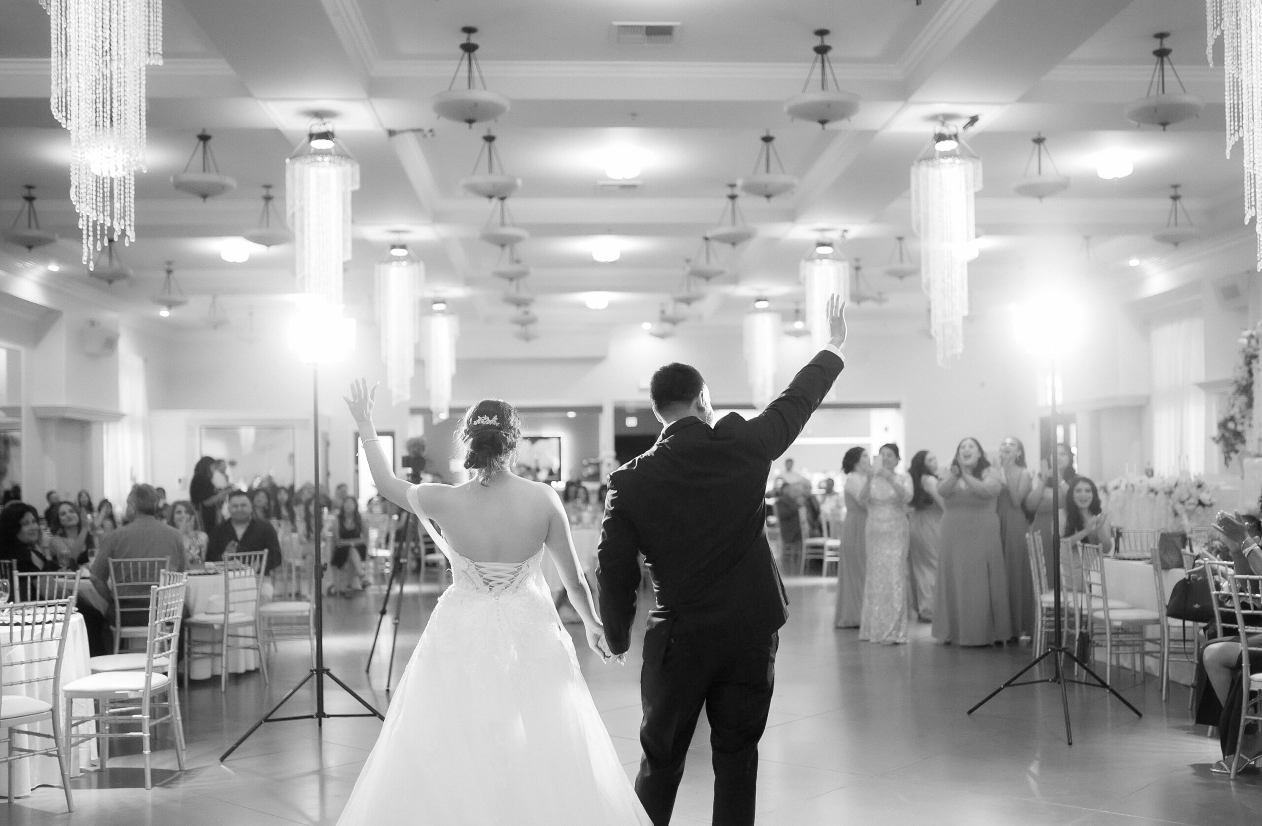 Bride and groom greet their guests after their first dance at an indoor wedding hall near me. Tall ceilings with grand chandeliers surround them.