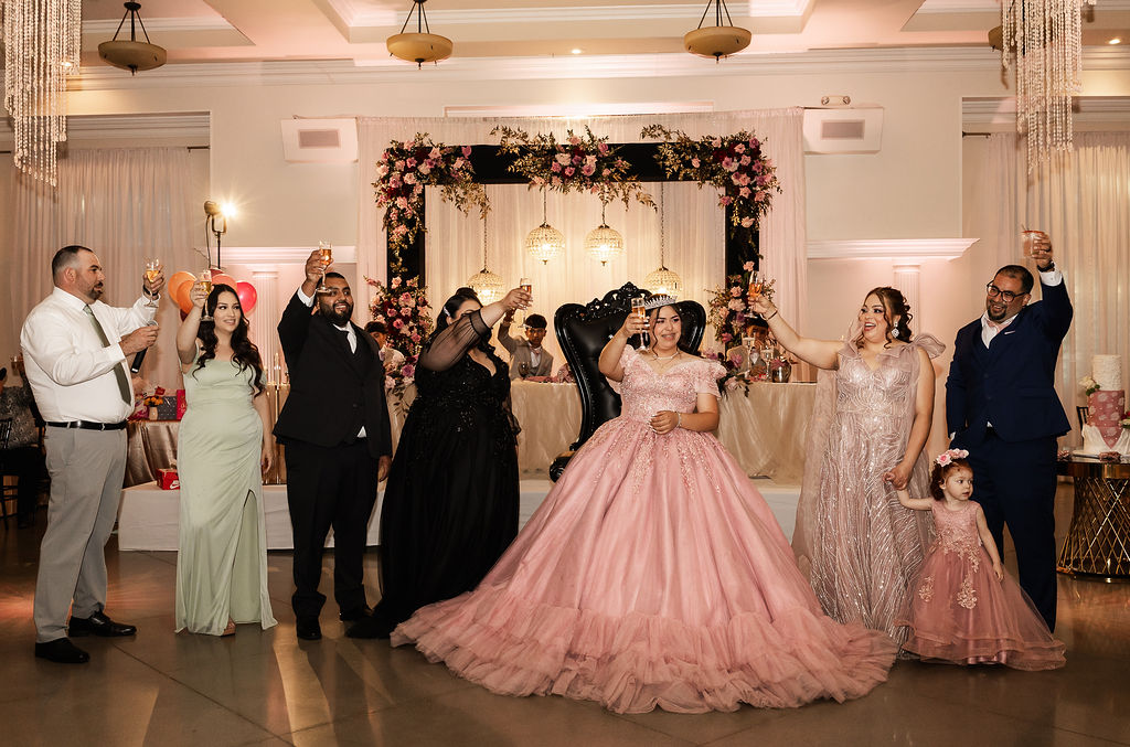 Pink and black Bakersfield quinceañera toasting with family in front of her main stage featuring luxury floral design and chandeliers. 