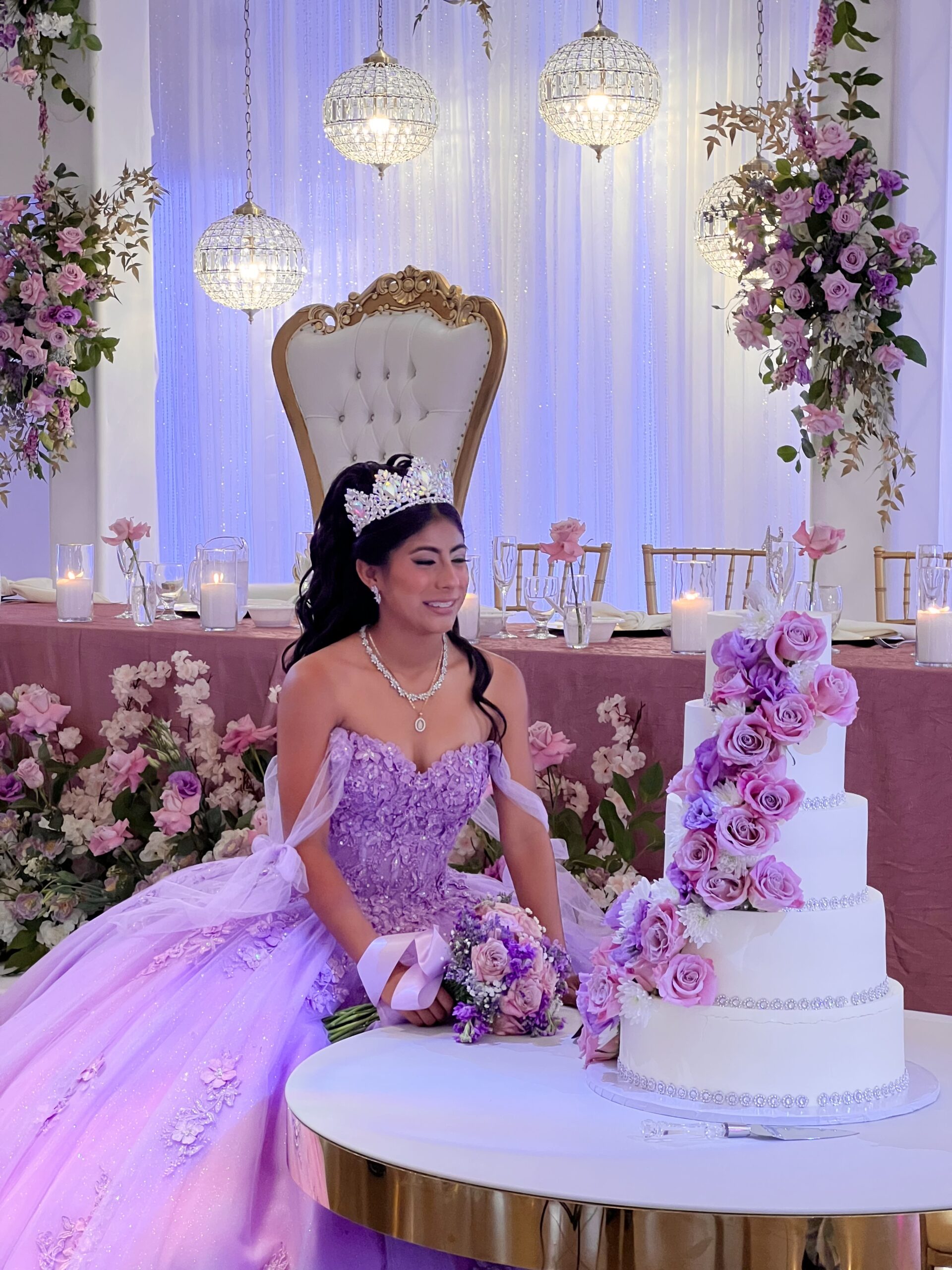 Quinceañera in a stunning quince dress posing in front of her main table and cake adorned with lilac flowers and shiny chandeliers