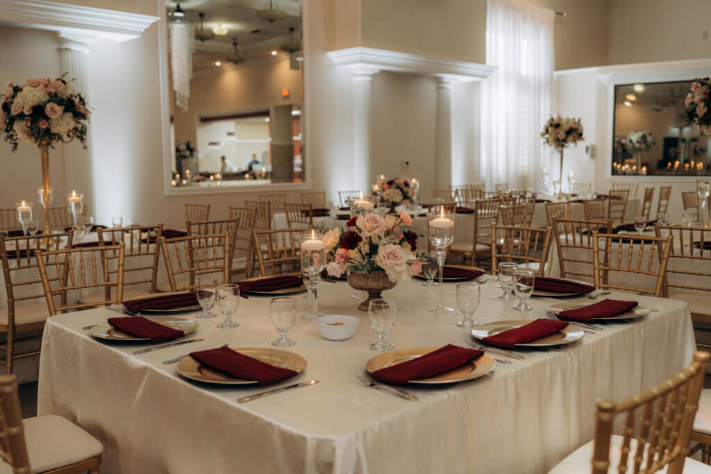 Traditional, elegant wedding and event venue in Bakersfield, California features columns, mirrors and chandeliers. The setup includes square guest tables with fresh floral featuring ivory, burgundy and blush for this candlelit dinner.
