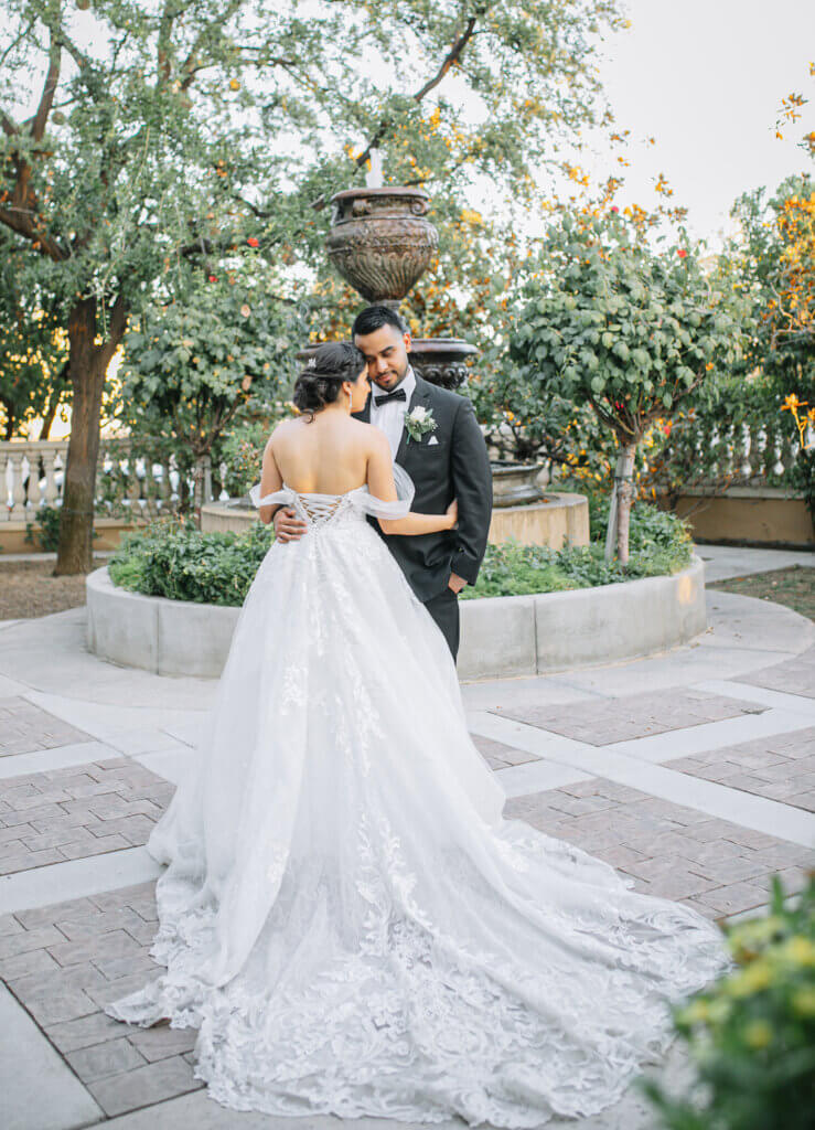 European style garden courtyard features a grand water fountain where a couple poses. Bride has on a dress with a long train with delicate lace details. 