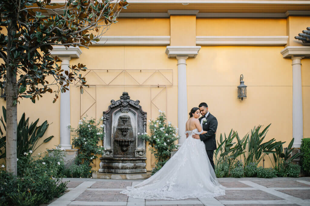 Bride and groom standing in front of a fountain surrounded by lush gardens in the garden courtyard of a Bakersfield Italian style wedding venue