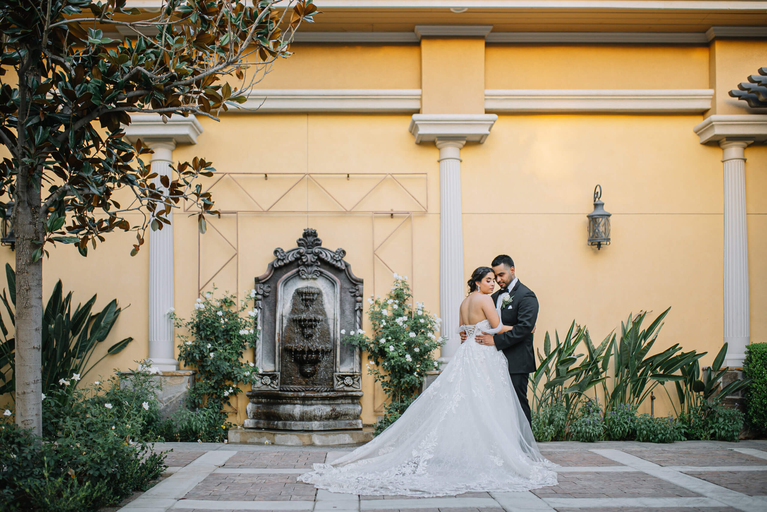Bride and groom standing in front of a wall fountain surrounded by lush gardens in the garden courtyard of a Bakersfield Italian style wedding venue