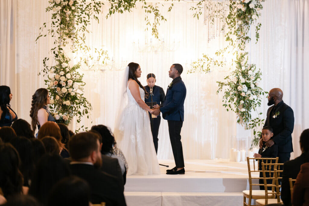 Bakersfield indoor wedding ceremony taking place in a reception hall. Bride is in a flowy wedding dress with floral appliqués and the groom is wearing a navy and black tuxedo standing in front of a custom floral statement piece with chandeliers.