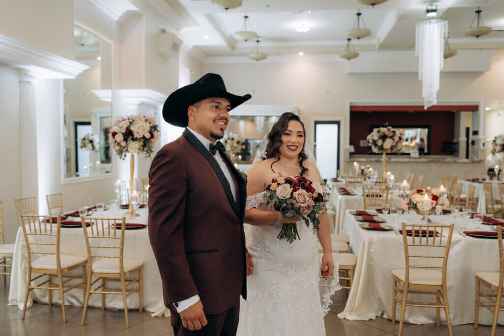 Just married couple doing a Bakersfield venue first the features tall and low centerpieces at the guest tables. Groom is a burgundy tuxedo and black hat while the bride has a beautiful lace dress and a bouquet of flowers featuring ivory, blush, and burgundy flowers.