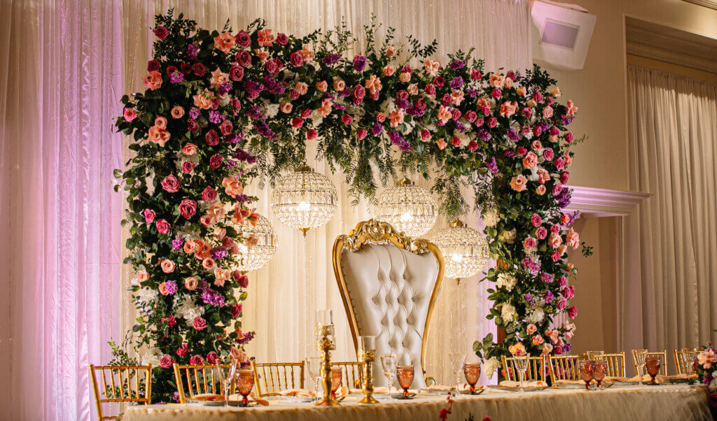 Bakersfield Luxury quinceañera head table featuring a floral filled arch in pinks, mauves, and lilacs, crystal chandeliers and a throne chair. 